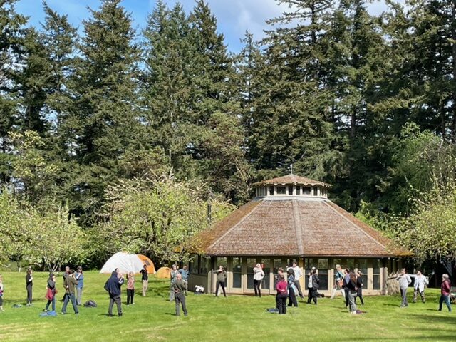 indralaya.pavilion A photo of a large pavilion in an apple orchard, with dozens of people standing outside it practicing mindful movement in the sunshine. Pine trees in the background and a hint of blue sky.