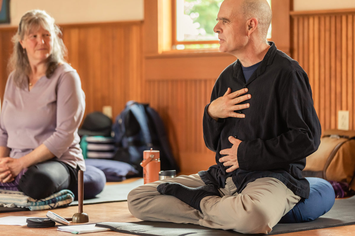 tim-raizelah-sitting-hands-mindfulness-kindness-retreat-wa-bellingham Tim Burnett, Lead instructor, sits with legs crossed and his hands on his torso as he explains something with deep feeling. Tim's wearing a black button down shirt and khakis,